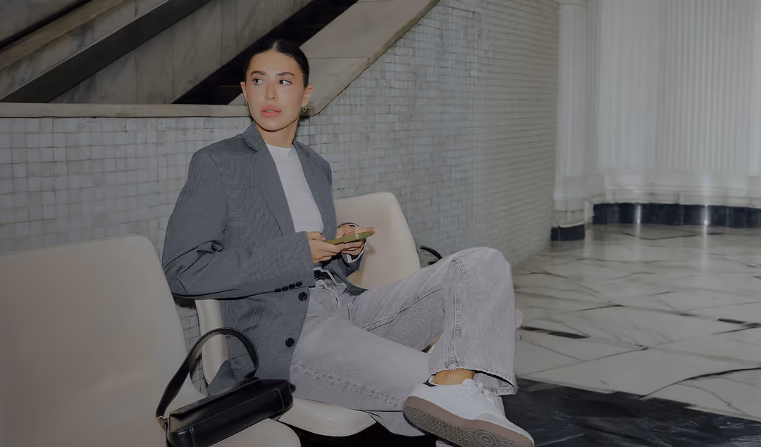 Donna con blazer grigio e jeans chiari seduta su una sedia bianca, con un telefono in mano e una borsa nera accanto.